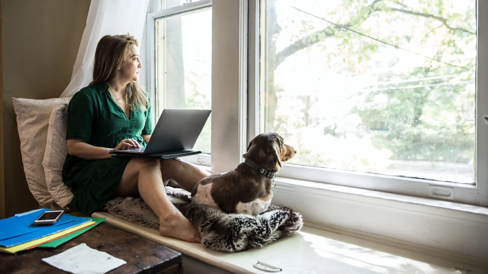 A woman sits in a reading nook in front of a window. On her lap is a laptop and to her feet is a cute little brown dog. She looks out of the window. So does her dog. 