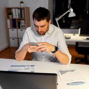 File image of a man looking at his phone in front of his laptop thumbnail
