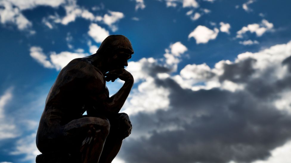  A bronze statue of a person deep in thought sits on a stone pedestal against a backdrop of stormy skies.