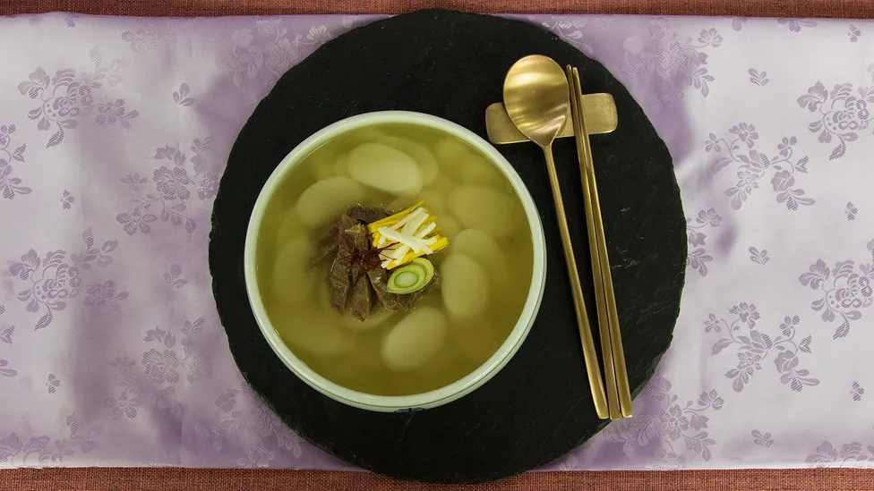 Bowl of soup containing round slices of Korean rice cake, beef, egg, and green onion, with golden chopsticks and spoon beside it.