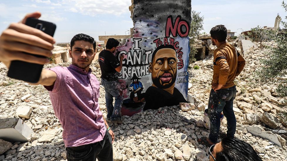 A friend of the muralist Aziz Asmar takes a selfie as the artist works on his Floyd memorial in Idlib, Syria (Credit: Izzeddin Idilbi/ Getty Images)