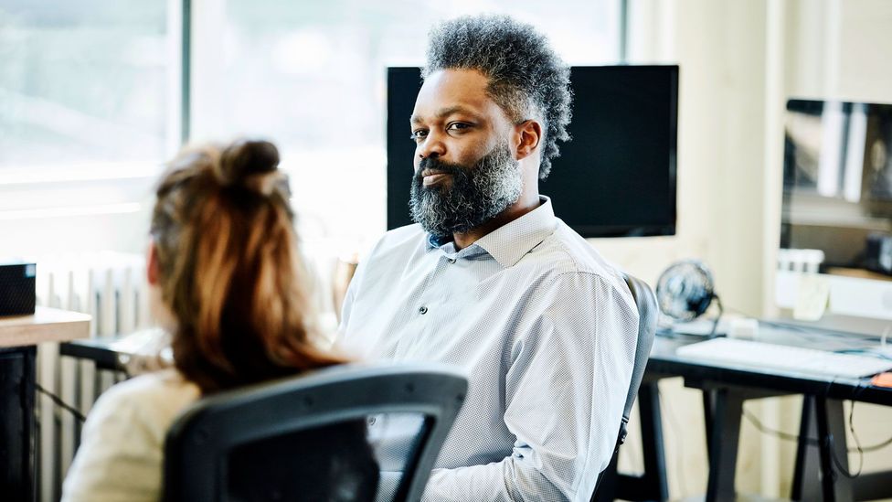 Man sits at desk chair talking to woman facing him