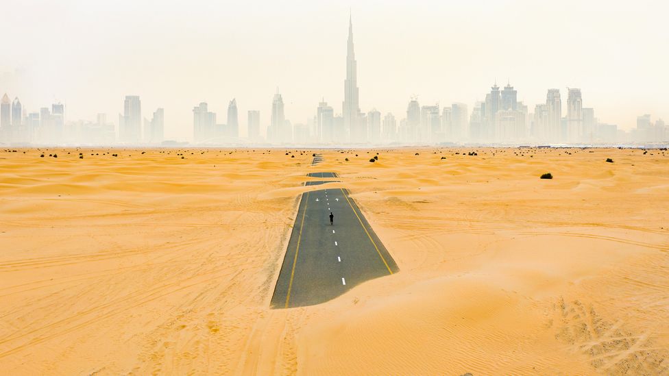 A person walks along a deserted road in the Dubai desert covered with sand dunes, towards a foggy Dubai skyscape (Credit: Travel Wild/ Alamy)