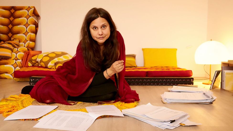 A portrait of Mithu Sanyal, a writer, surrounded by manuscripts (Credit: Carolin Windel)