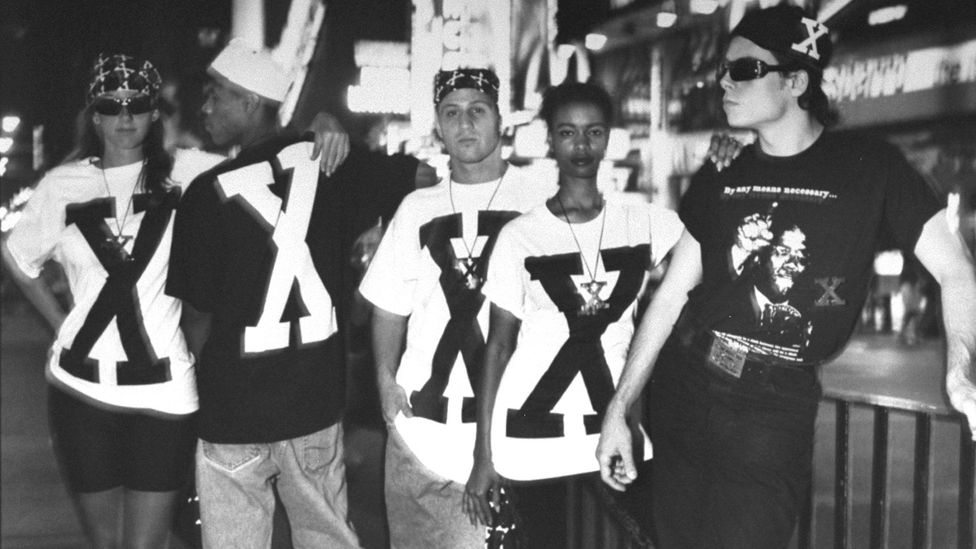Group of men & women wearing 'X' emblazoned t-shirts, bandanas, baseball caps, and leather necklaces, with one man clad in a Malcolm X t-shirt