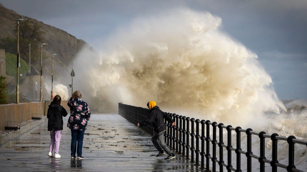 Waves crash over a walkway on the coast as people look on (Credit: Getty Images)