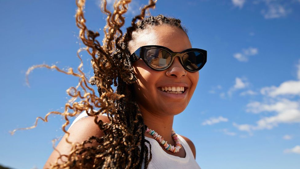 Woman in sunglasses, a necklace and a white vest smiles (Credit: Getty Images)