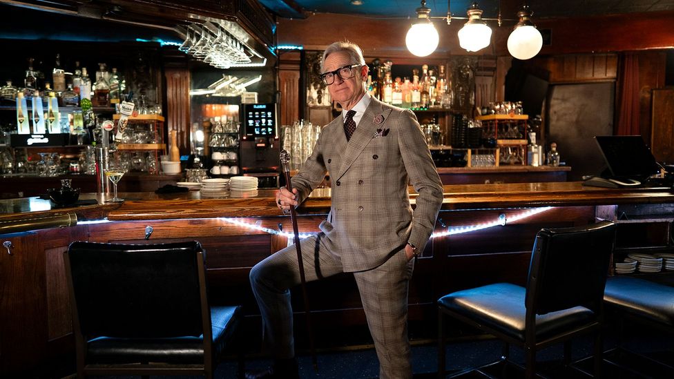 Paul Feig stood in front of the bar next to bar stools in a cocktail bar, wearing a grey suit and holding a cane (Credit: Ryan Silver)