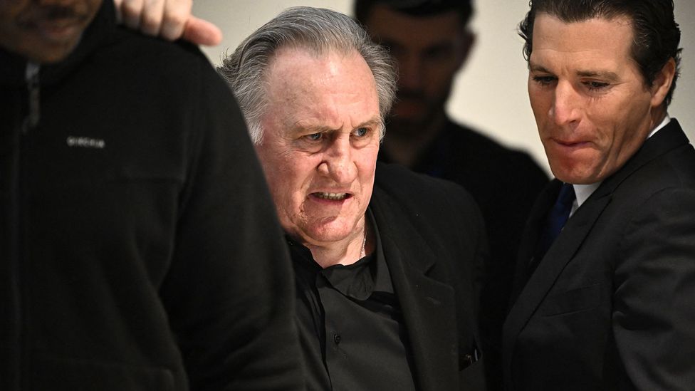 Gérard Depardieu wearing a black suit with a policeman behind him (Credit: Getty Images)