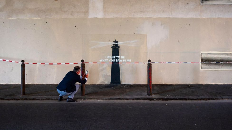 A man taking a photo of Banksy's new mural Lighthouse in Marseille, with text that reads 