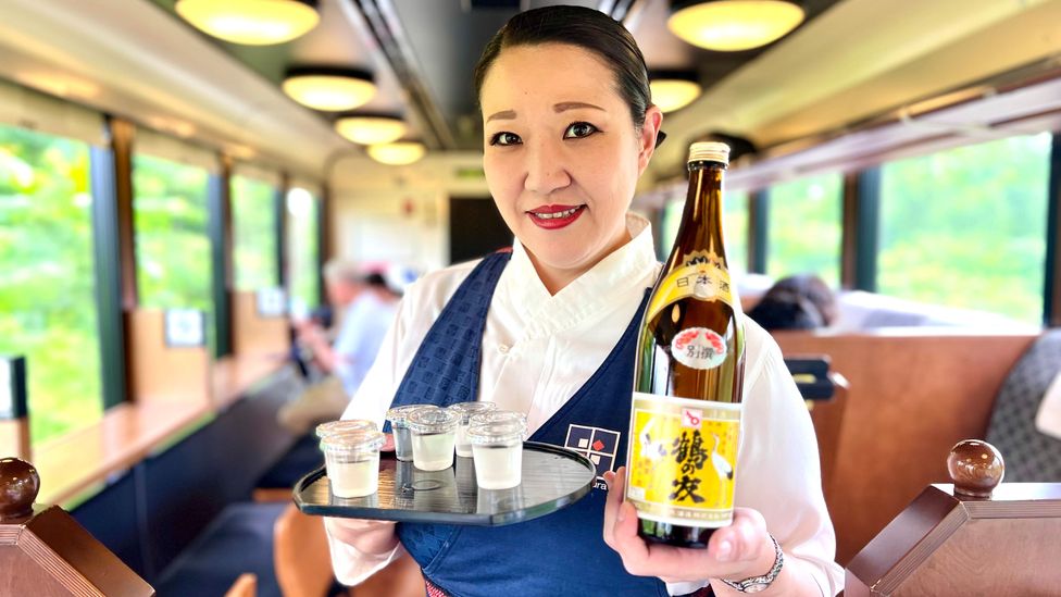 Close up of female server holding a bottle of sake and glasses on a tray on Japan's Shu*Kura sake train (Credit: Kitty Knowles)
