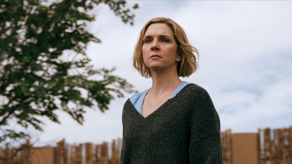 A still from Pluribus showing Rhea Seehorn, a woman with a blonde bob, wearing a green v-neck jumper and blue t-shirt, standing outside (Credit: Apple TV+)