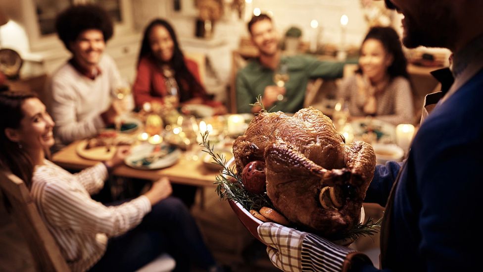 A man carrying a roasted turkey to a table of friends sat at a table for Thanks Giving (Credit: Getty Images)