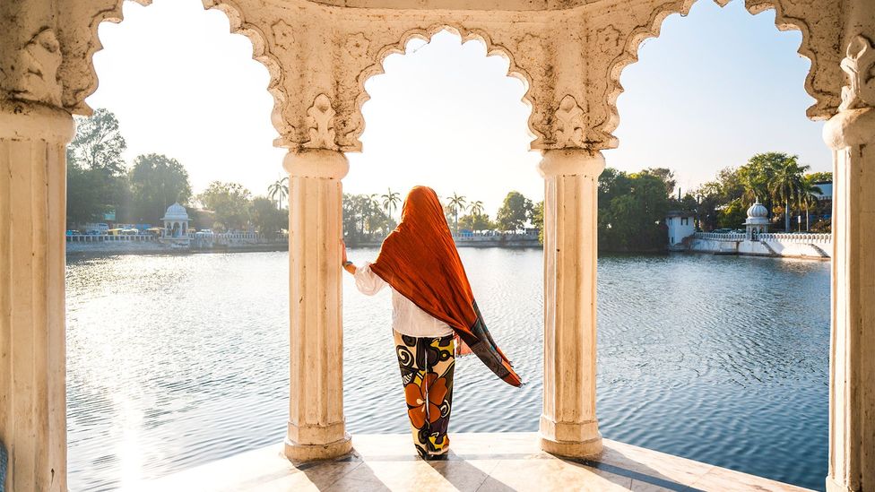 Woman with orange headscarf stands between carved stone arches looking out onto a tranquil lake in Udaipur (Credit: Getty Images)