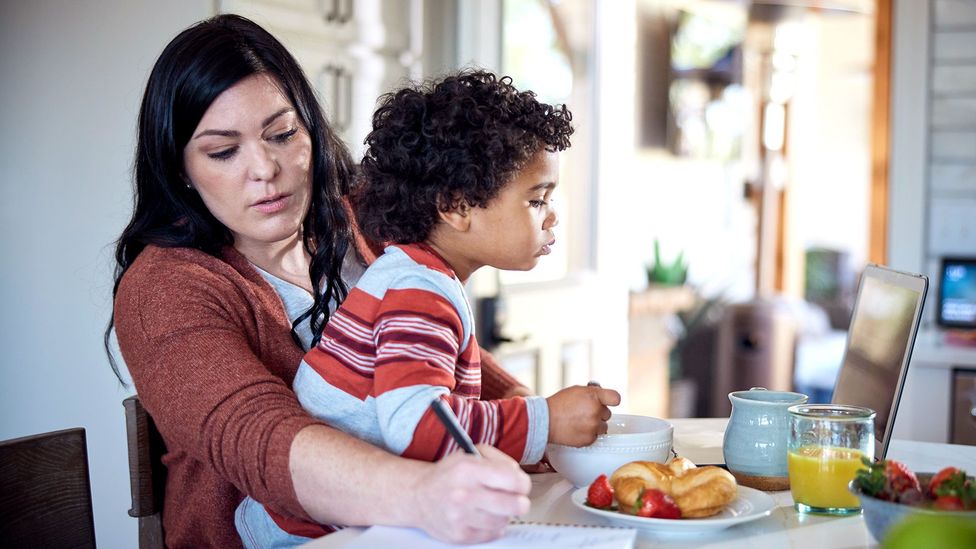 A mother holding a child on her lap as he eats breakfast and she writes on a paper (Credit: Getty Images)