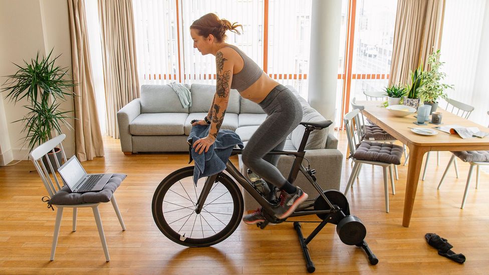 A woman pedalling on an exercise bike in her living room with a laptop on a chair in front of her (Credit: Getty Images)