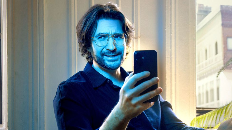 The author Thomas Germain sits holding a smartphone, with blue light shining on him (Credit: Hana Mendel)