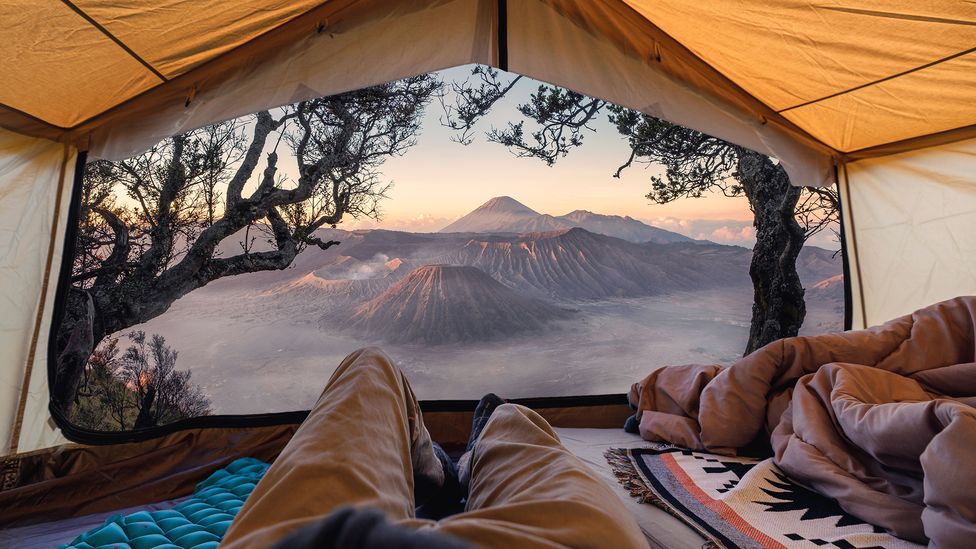 Two people lie on sleeping mats in a tent overlooking a mountainous view, with two trees framing the tent canopy in the foreground (Credit: Getty Images)