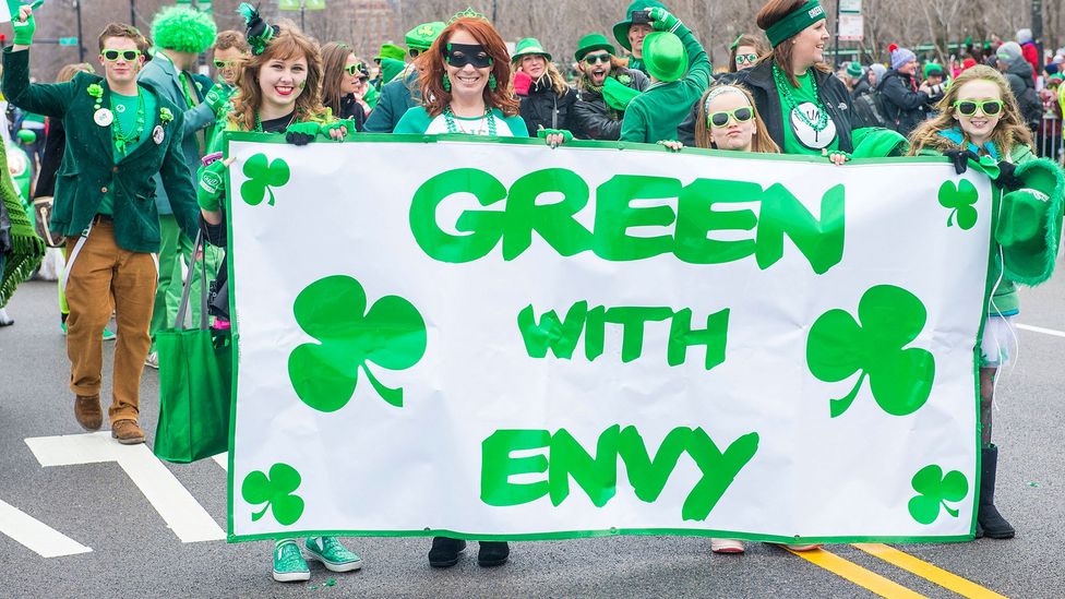 At a parade, four young women dressed in green hold a banner that reads "Green with Envy", with six clovers spaced around the words (Credit: Alamy)