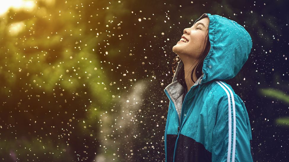 A woman in a green sports jacket with the hood up smiles as she leans her head up, eyes closed, towards the falling rain (Credit: Getty Images)