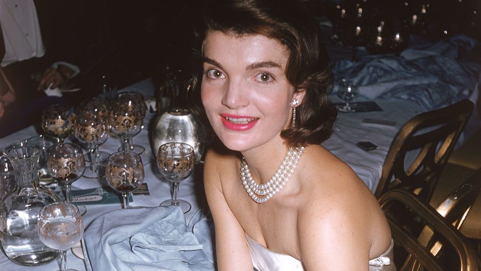 Jacqueline Kennedy Onassis poses for a photo sitting in a white dress and pearl necklace, with wine glasses on a white-tablecloth table behind her (Credit: Getty Images)