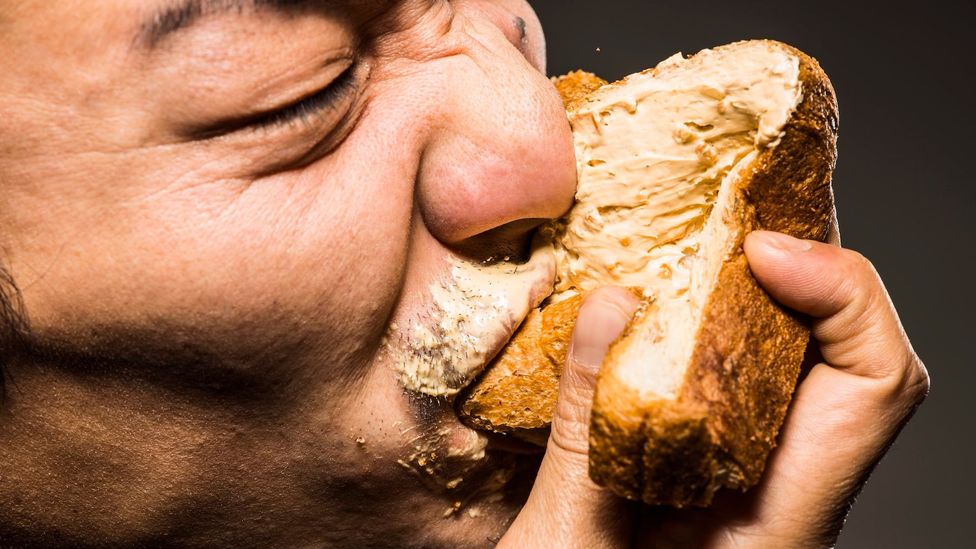 A close-up of a person stuffing their face with a bread-like food covered in beige spread (Credit: Getty Images)