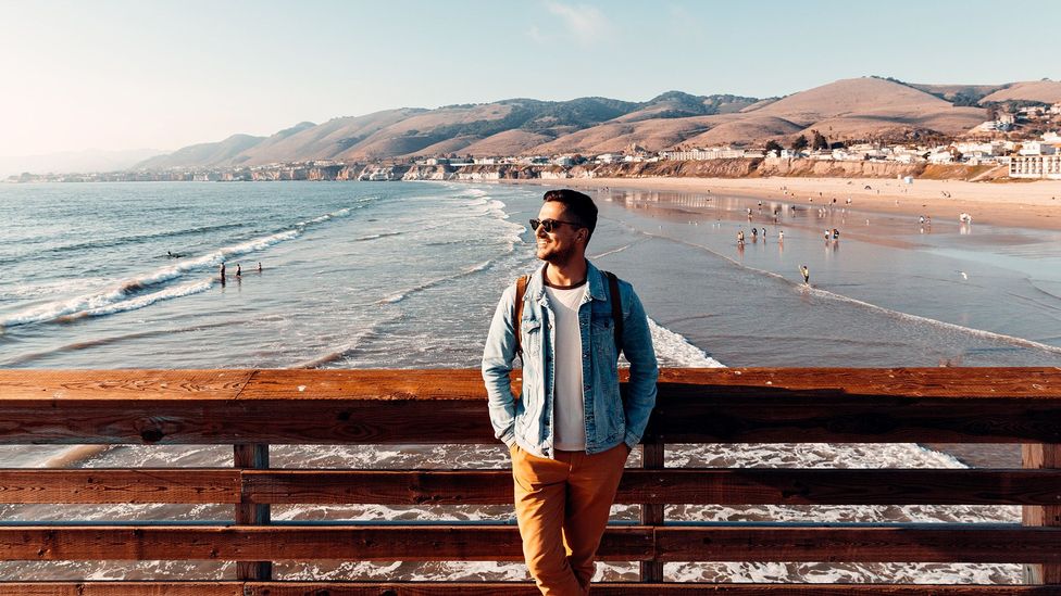 Man leaning against pier fence with beach and coastline behind him at Pismo Beach, California (Credit: Getty Images)