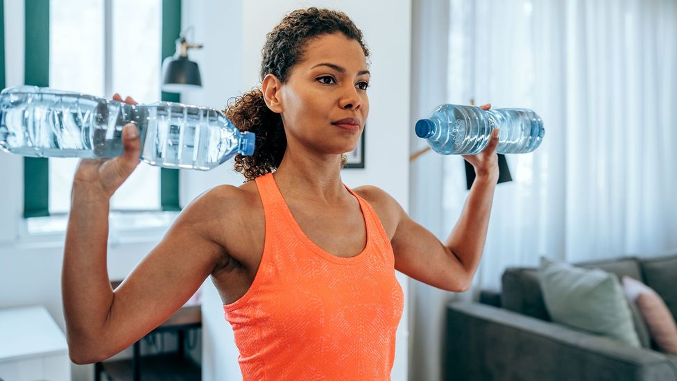 A woman in an orange sports top with a water bottle in each hand performing a shoulder press (Credit: Getty Images)