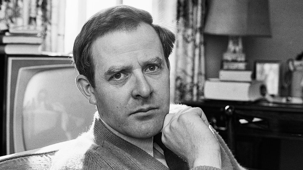 A black and white portrait of John John le Carré sat in a living room, with a television behind him (Credit: Getty Images)