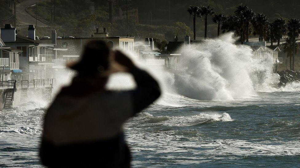 A man wearing a hat looks at stormy waves crashing over a balustrade, with palm trees and houses in the background, during a strong El Niño event in California in 2016 (Credit: Getty Images)