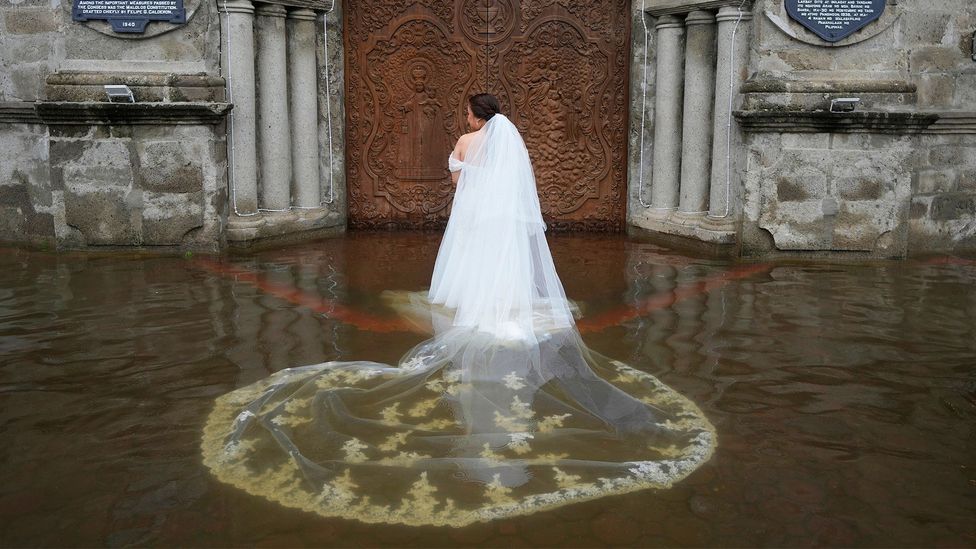 A woman wearing a wedding dress and a soaked veil stands, with her back to the camera, in brown floodwater in front of a large wooden church door in the Philippines (Credit: Aaron Favila/ Associated Press)