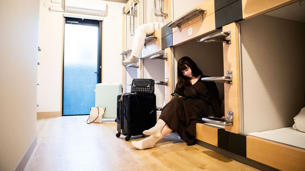 A traveller sits beside her luggage in a hostel dormitory with capsule style bunks (Credit: Getty Images)